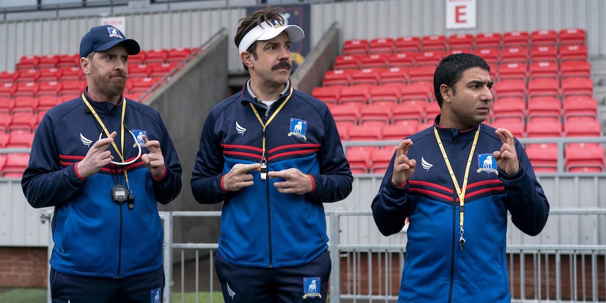 Nate, Ted Lasso and Coach Bird cross their fingers on the field