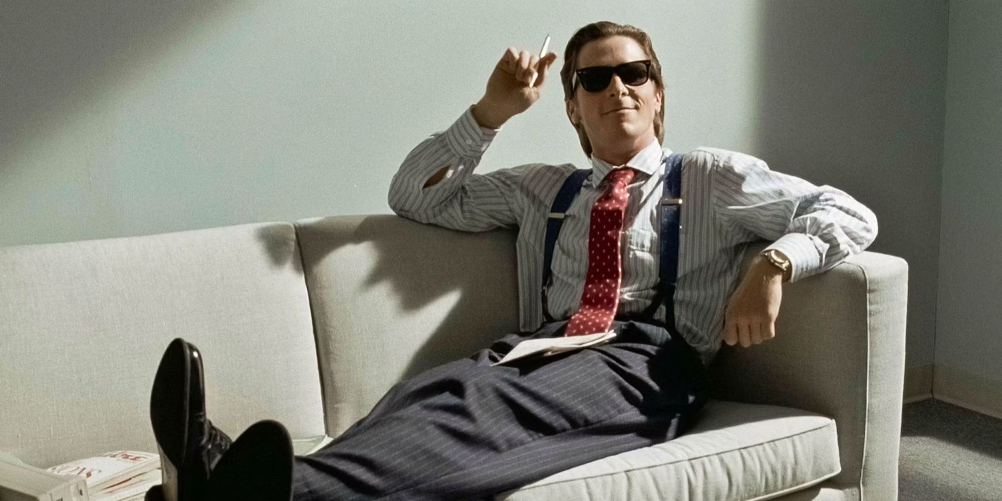 Patrick Bateman (Christian Bale) smokes on the couch and wears sunglasses in American Psycho.