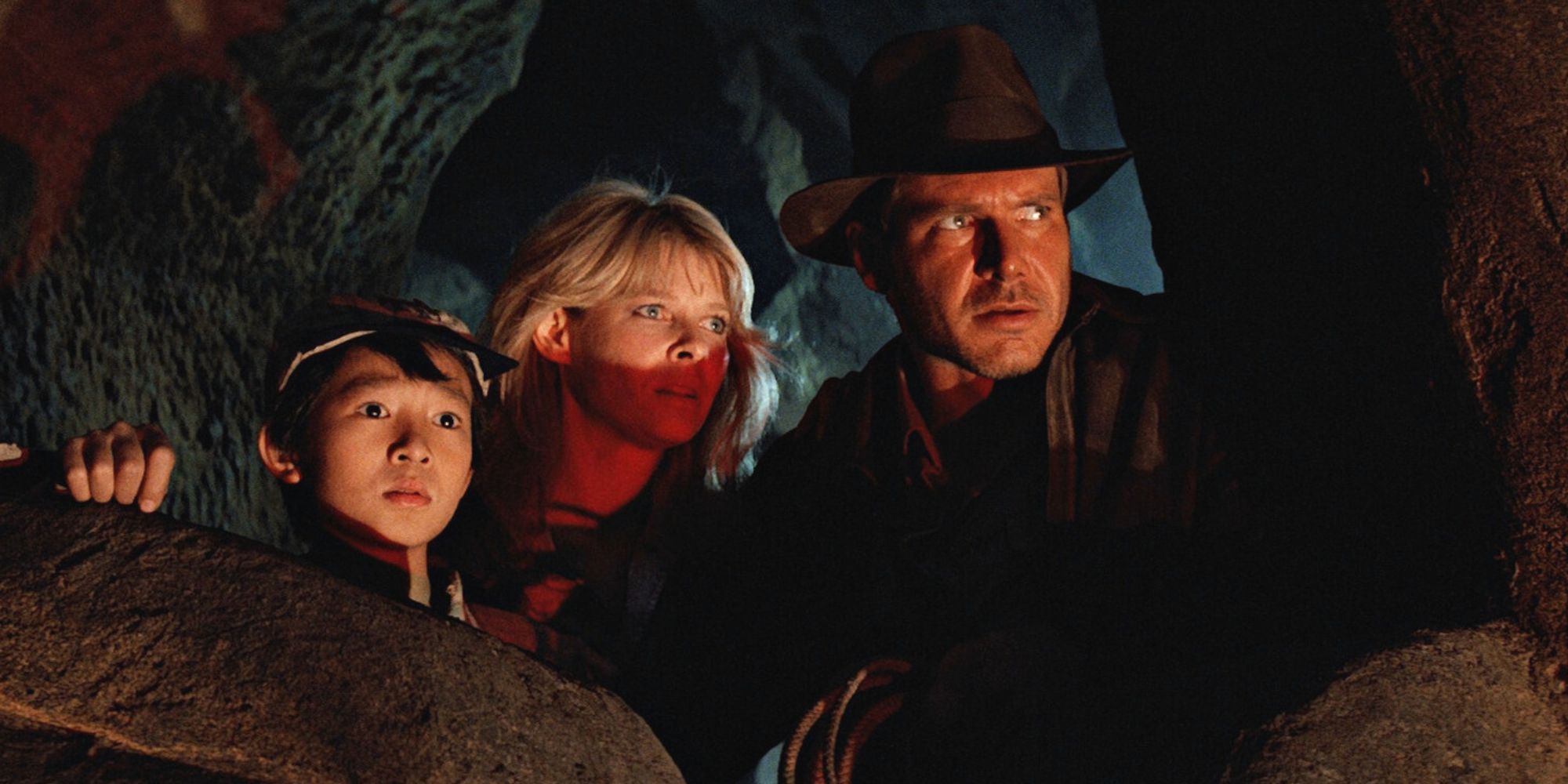 Ke Hai Quan, Kate Capshaw and Harrison Ford crouch behind rocks in a cave, looking up slightly in Indiana Jones and the Temple of Doom.