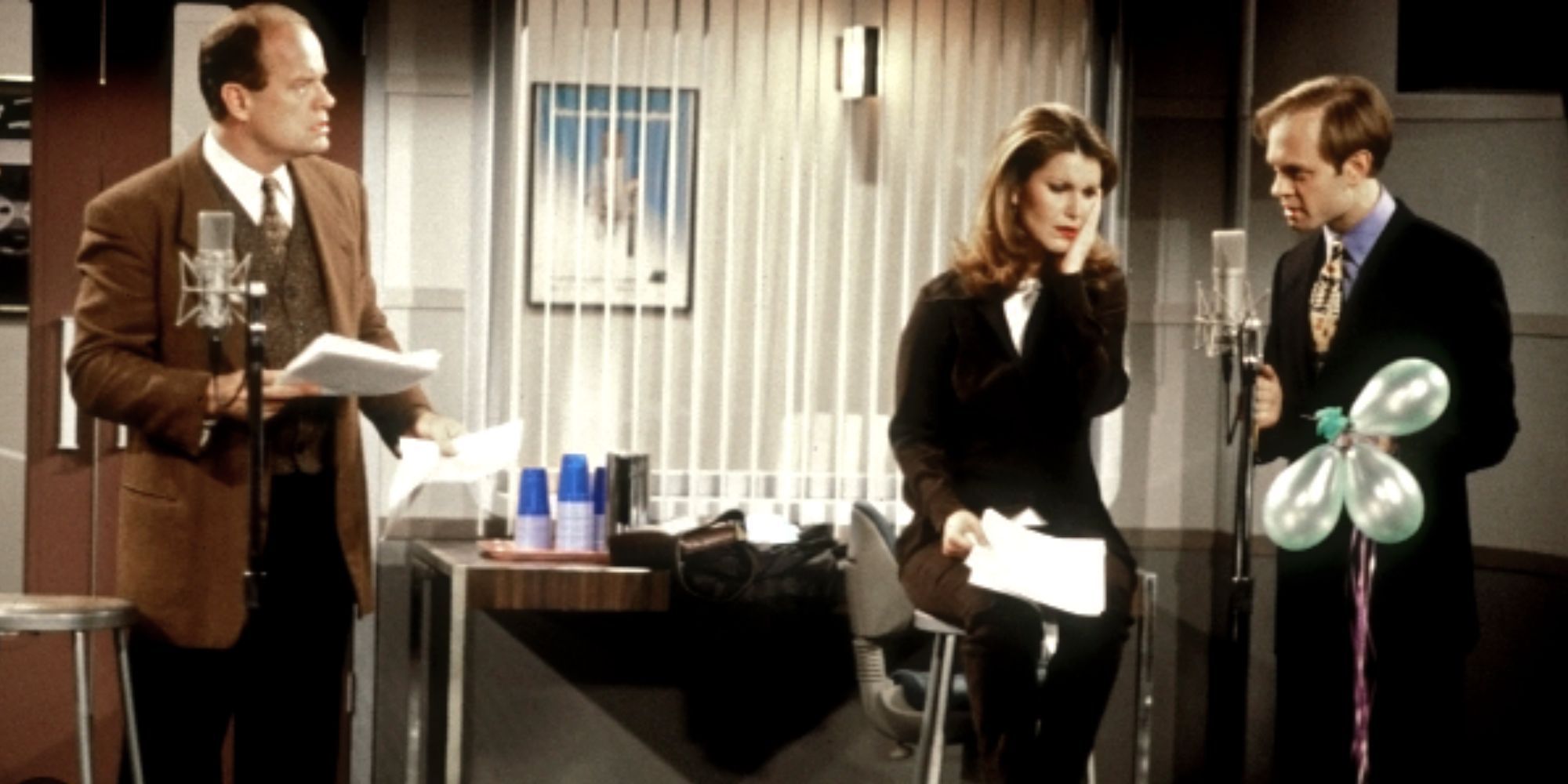 Kelsey Grammer stands in front of a microphone and looks at Peri Gilpin and David Hyde Pierce next to him in Fraser.