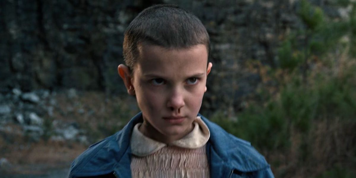 Millie Bobby Brown as Eleven wears a pink dress with a blue jacket.  She looks straight ahead, using her telekinetic abilities.