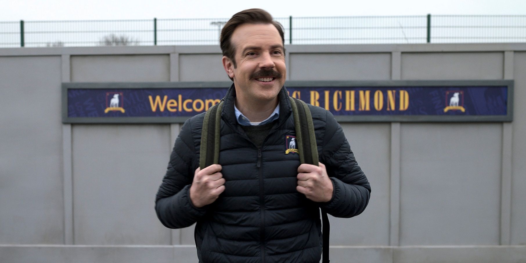 Jason Sudeikis smiling with a backpack on the soccer field in Ted Lasso.