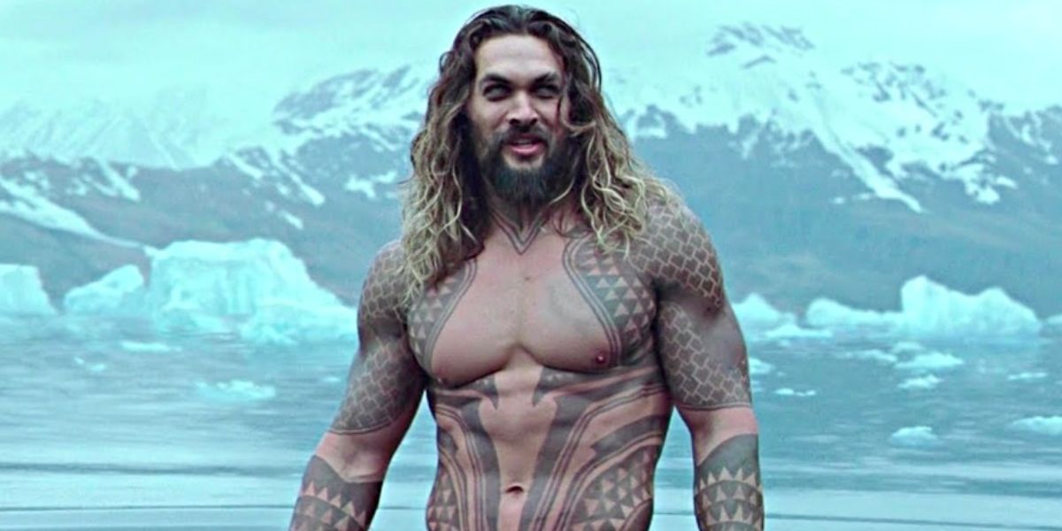 Jason Momoa as Aquaman stands shirtless in the water in front of the mountains.