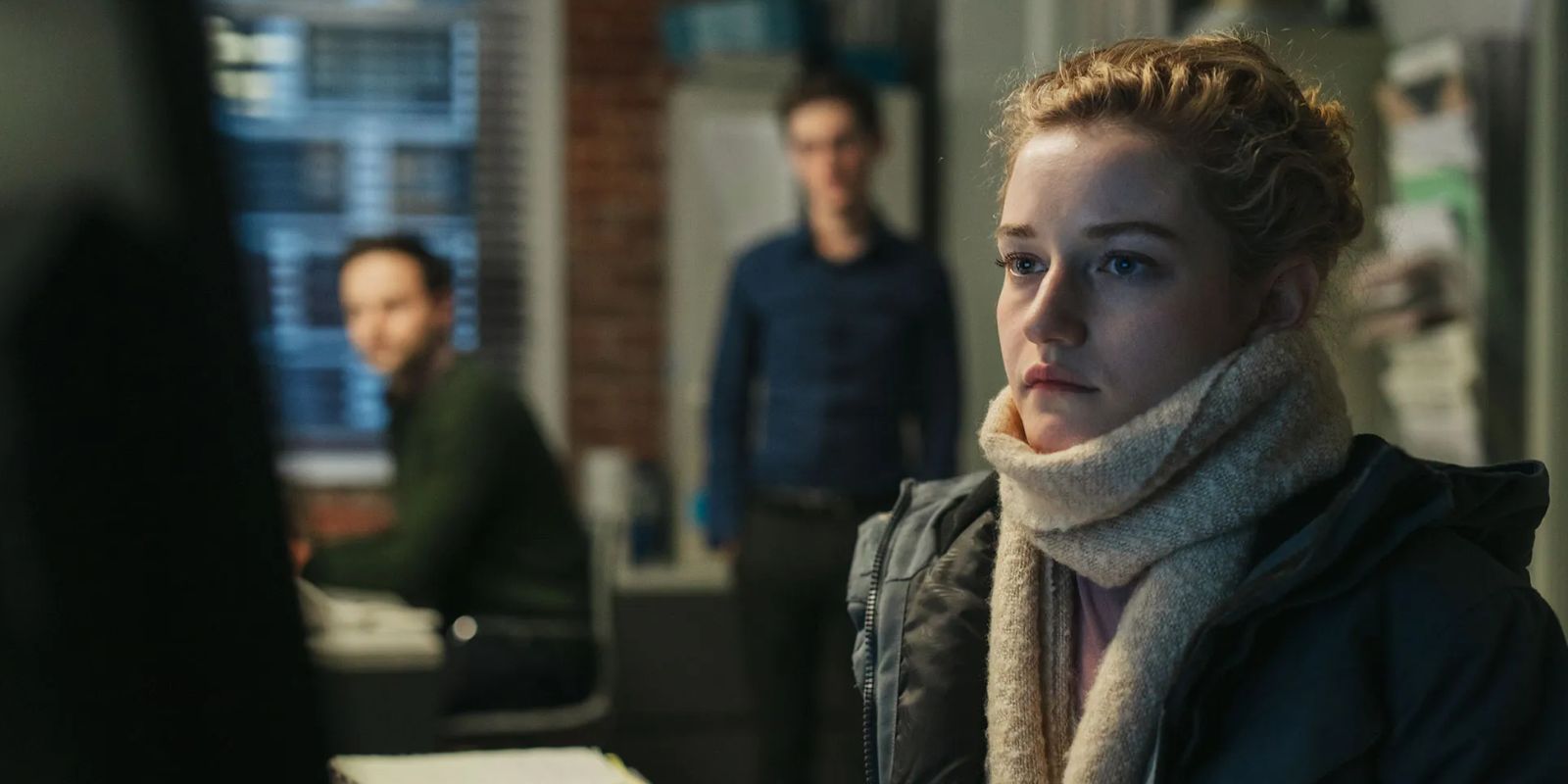 A woman sits at a table as two men loom behind her and watch her work.